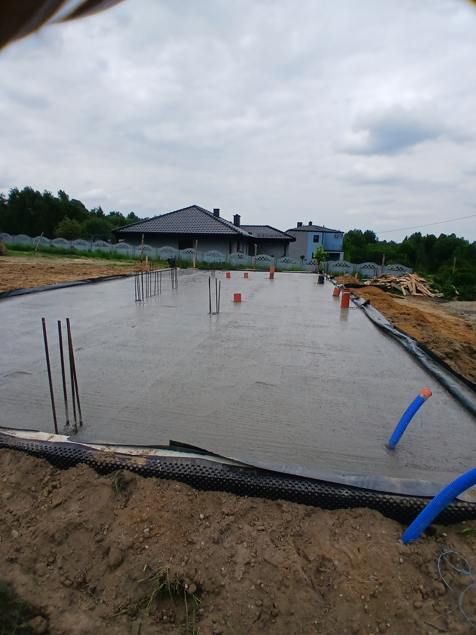 Wylana płyta fundamentowa z wystającymi prętami zbrojeniowymi i rurami instalacyjnymi. Widok na budynek w tle, pochmurne niebo.