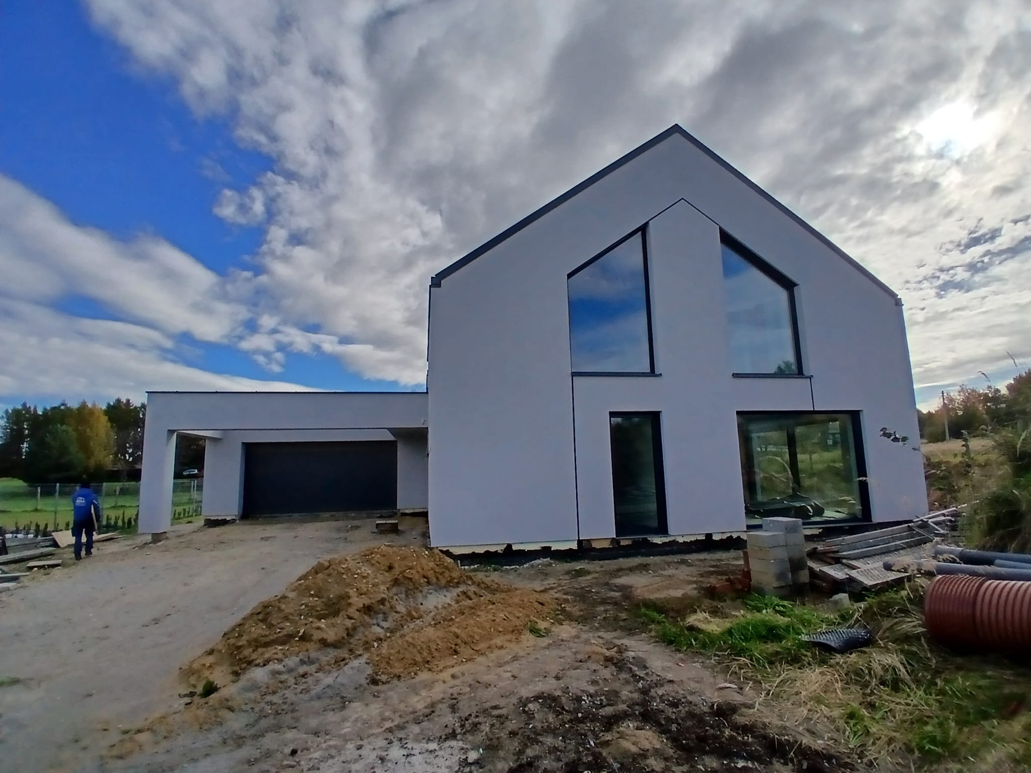 Nowoczesny dom z szarą elewacją i dużymi oknami, widoczny z zewnątrz. Na pierwszym planie ziemia i materiały budowlane, w tle błękitne niebo z chmurami.