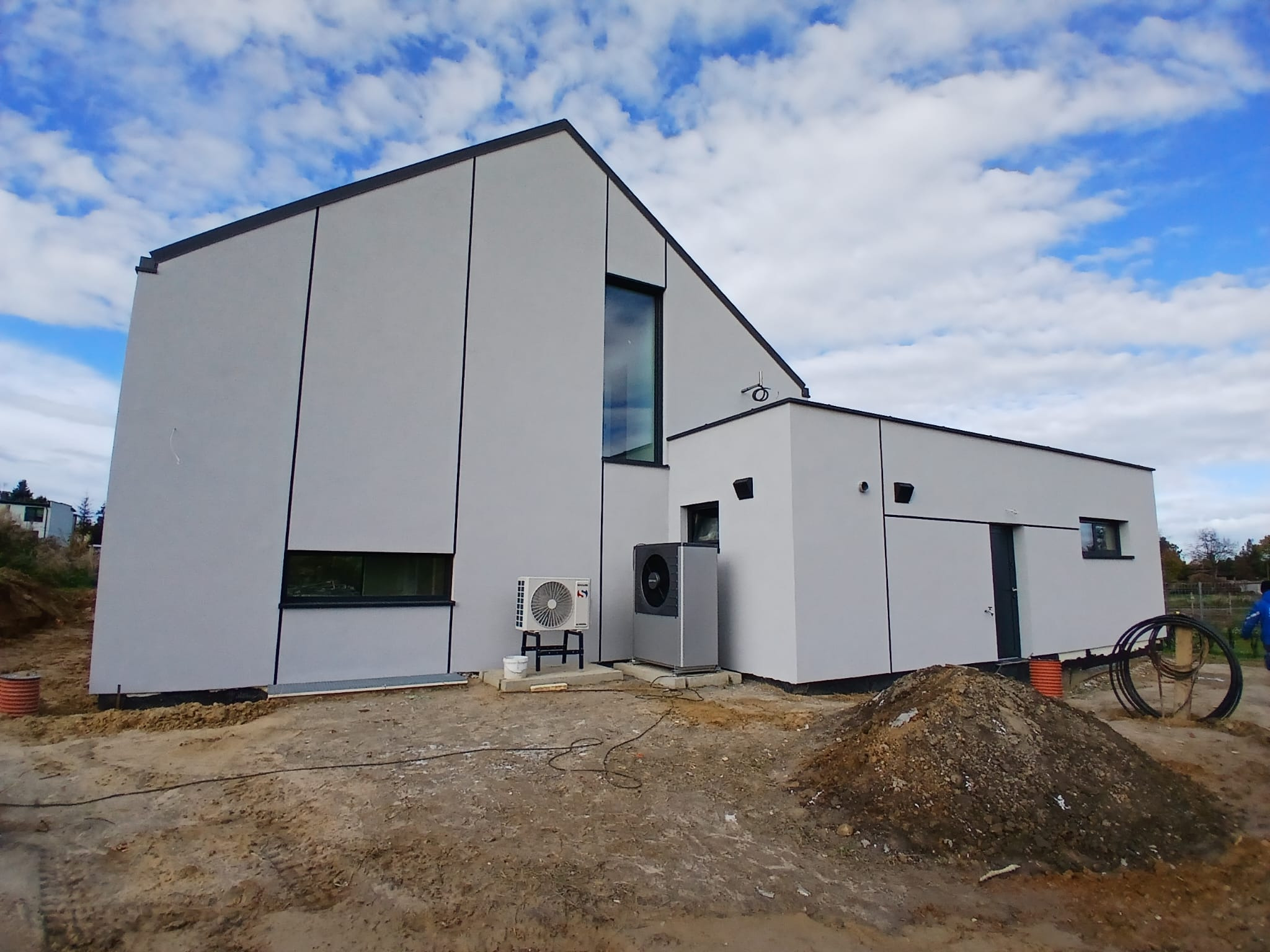 Nowoczesny dom z szarą elewacją i czarnymi akcentami, widoczny z boku. Na zewnątrz pompa ciepła i teren w trakcie prac ziemnych. Błękitne niebo z chmurami w tle.