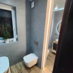 Nowoczesna łazienka z szarą glazurą, drewnopodobną podłogą, podświetlanym lustrem i zabudową WC typu Geberit.