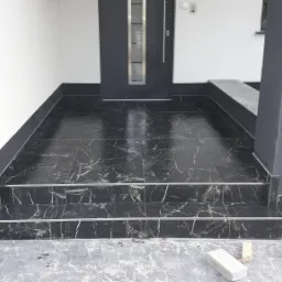 Wykończone czarnym marmurem schody wejściowe do domu w Trzcinicy, z widocznymi srebrnymi listwami wykończeniowymi i fragmentem elewacji.