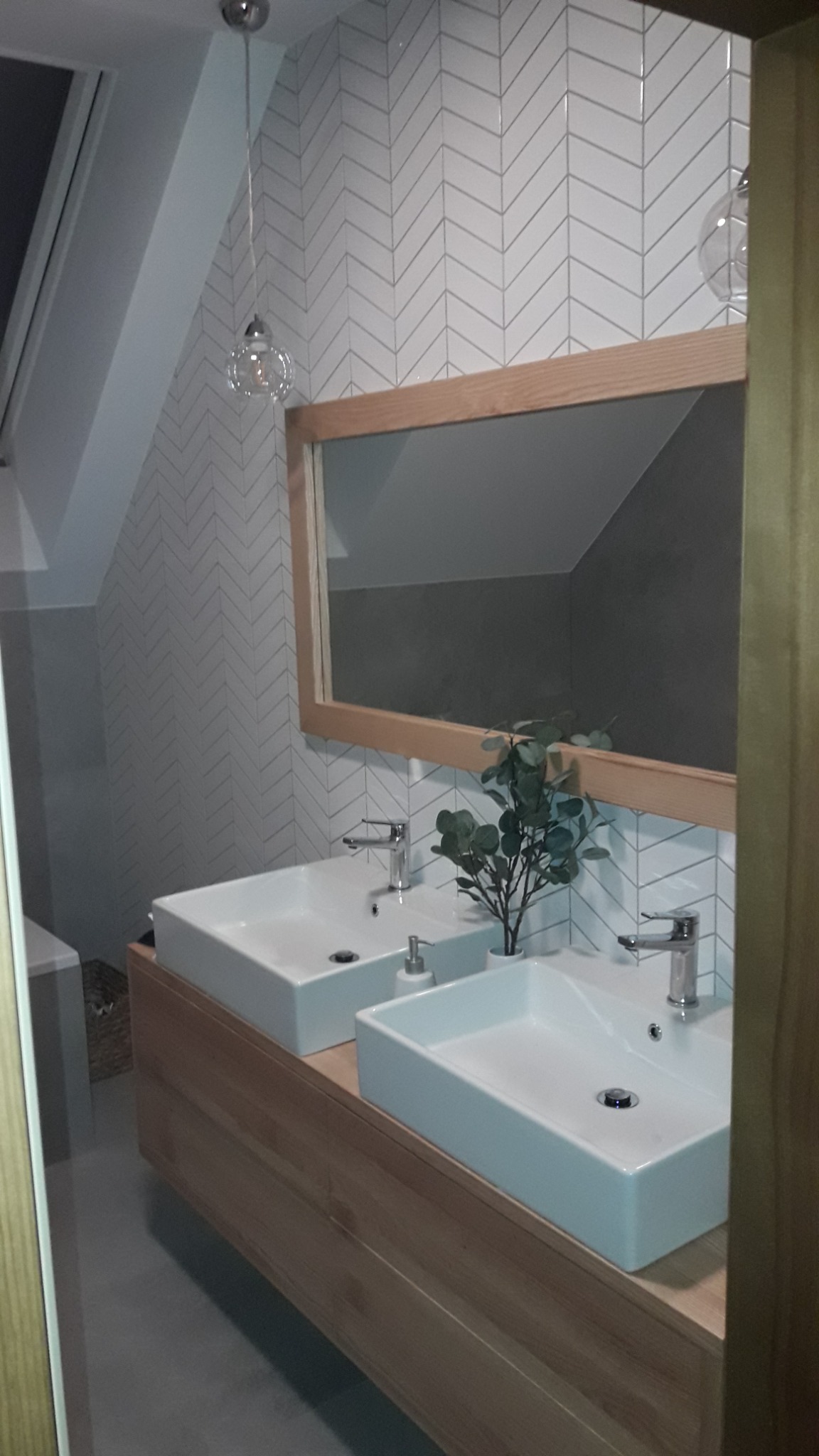 Nowoczesna łazienka na poddaszu z dwoma białymi umywalkami nablatowymi, drewnianą szafką pod umywalkami, lustrem w drewnianej ramie, białymi płytkami ułożonymi w jodełkę na ścianach oraz wiszącymi...