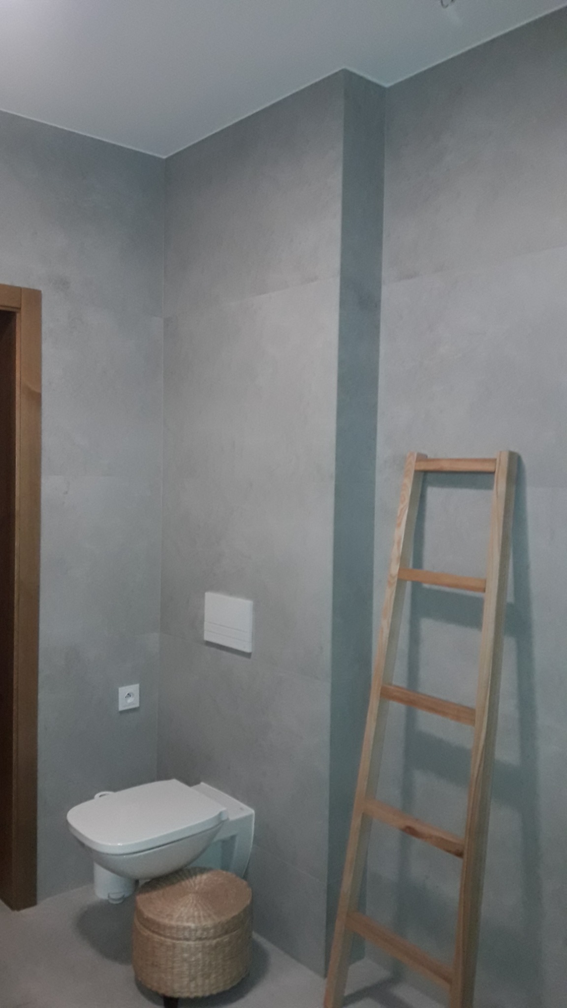 Wykończona łazienka w szarościach z podtynkową toaletą, dekoracyjną drabinką drewnianą i koszem z wikliny, widoczne szare płytki imitujące beton.