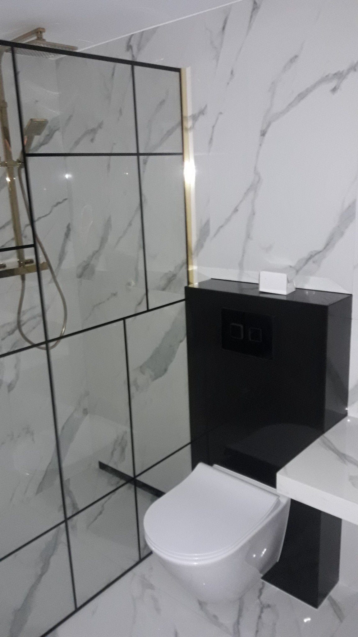 Nowoczesna łazienka z kabiną prysznicową typu walk-in z czarnymi profilami, podwieszanym sedesem z czarną zabudową i marmurowymi płytkami na ścianach i podłodze.