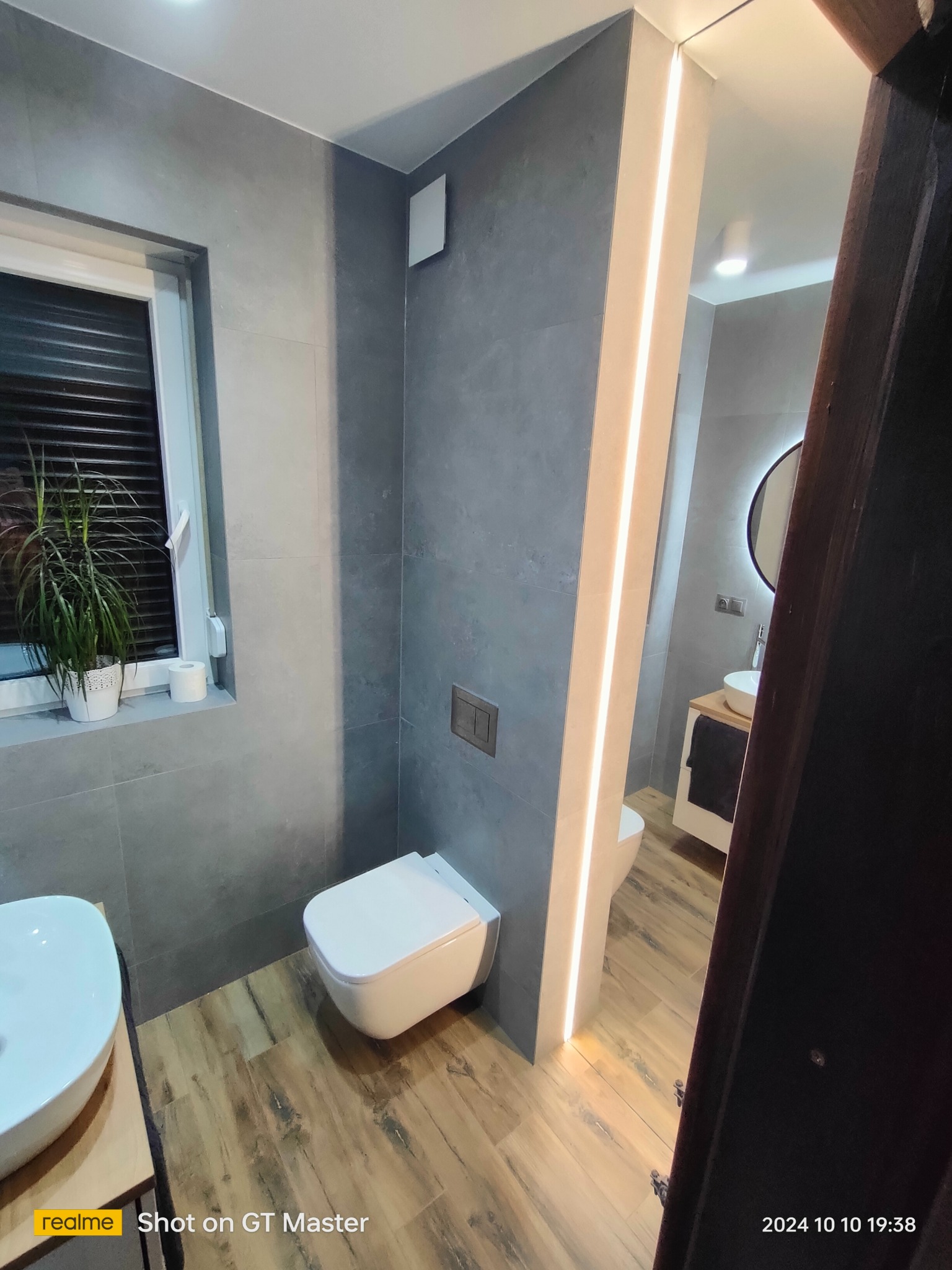 Nowoczesna łazienka z szarą glazurą, drewnopodobną podłogą, podświetlanym lustrem i zabudową WC typu Geberit.