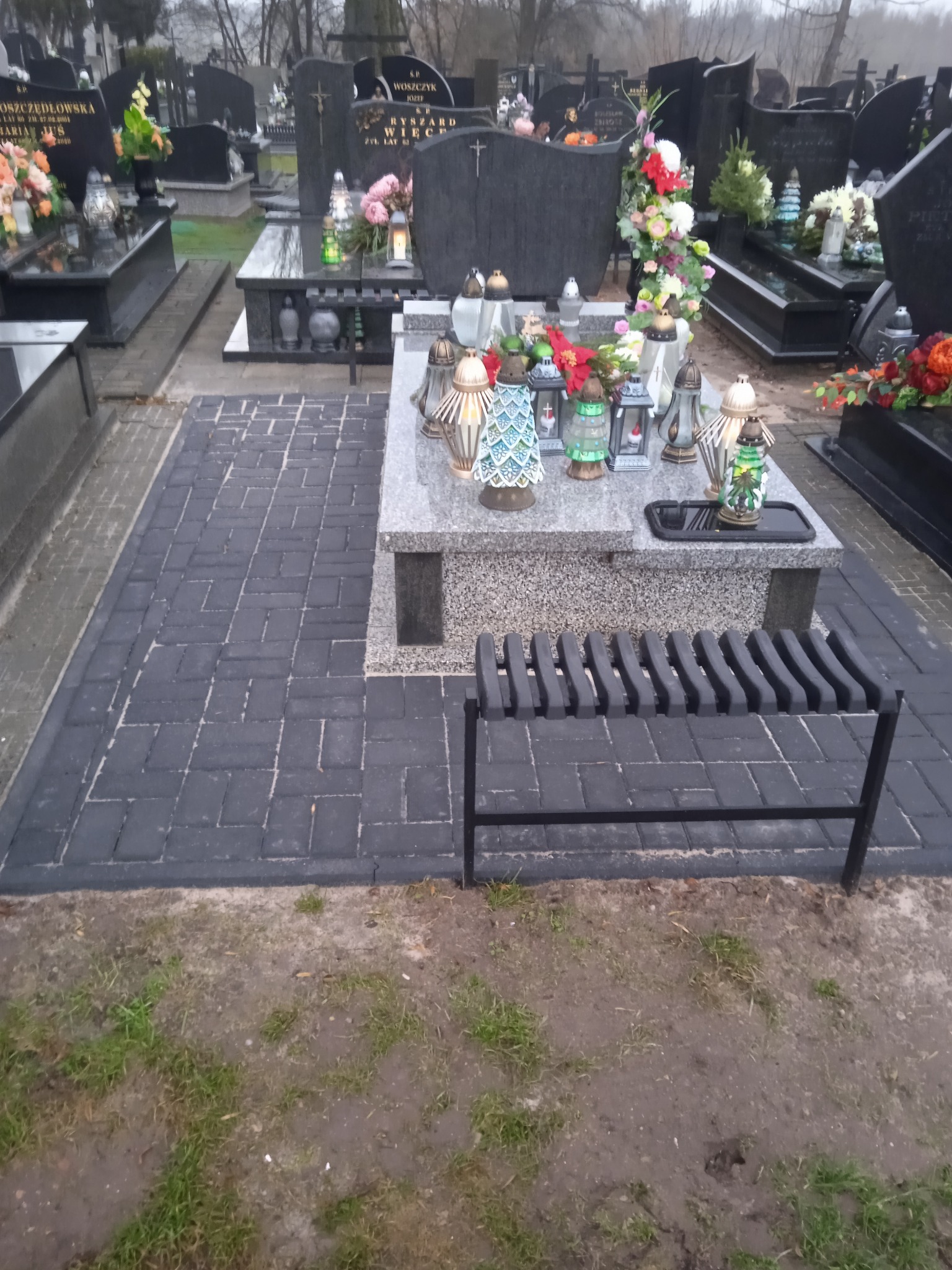 Ciemnoszara kostka brukowa ułożona wokół nagrobka z ławką na cmentarzu, widoczne znicze i kwiaty.