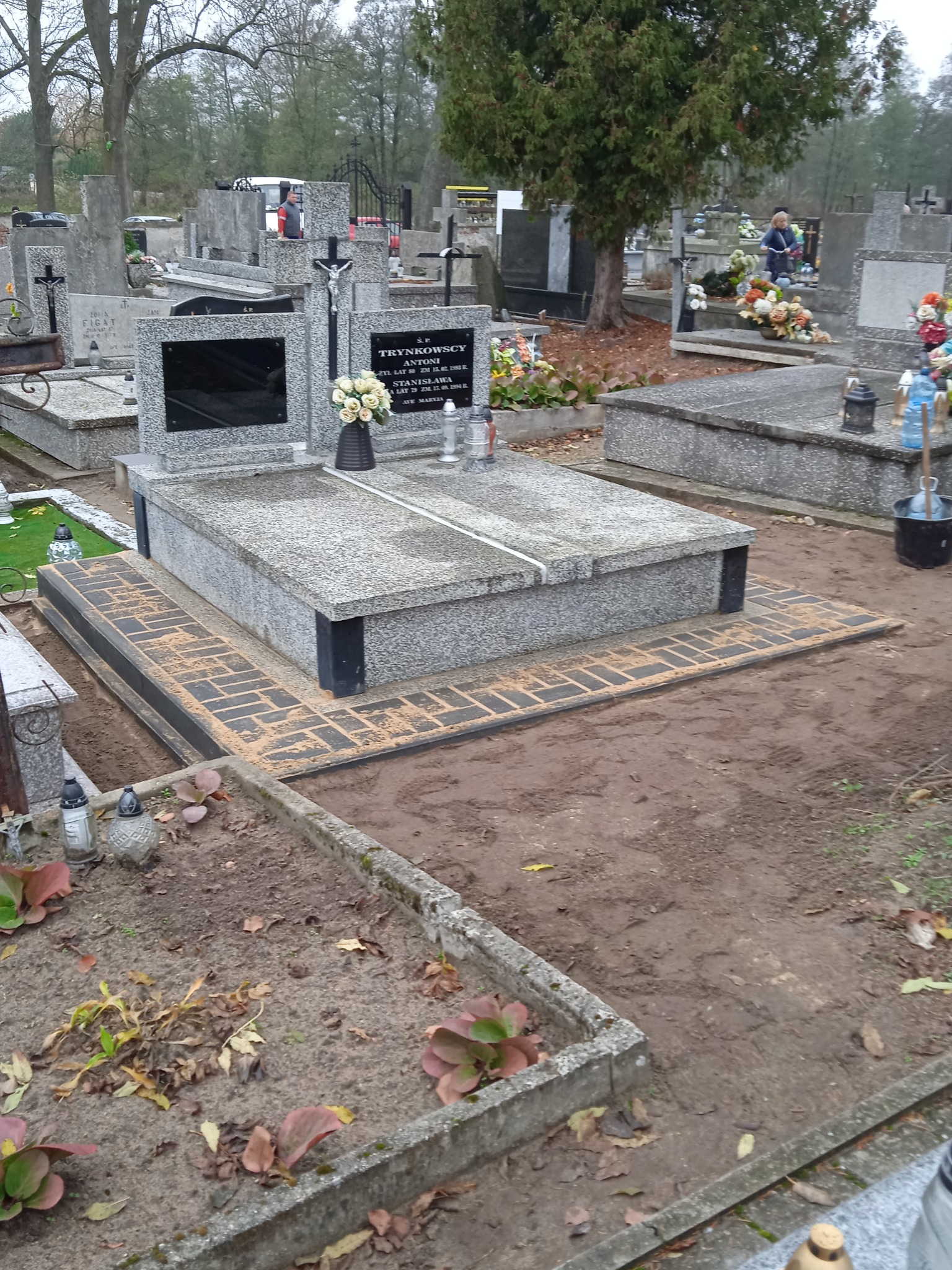 Granitowy nagrobek otoczony nową, ciemną kostką brukową ułożoną w cegiełkę, na cmentarzu z jesiennym krajobrazem w tle.