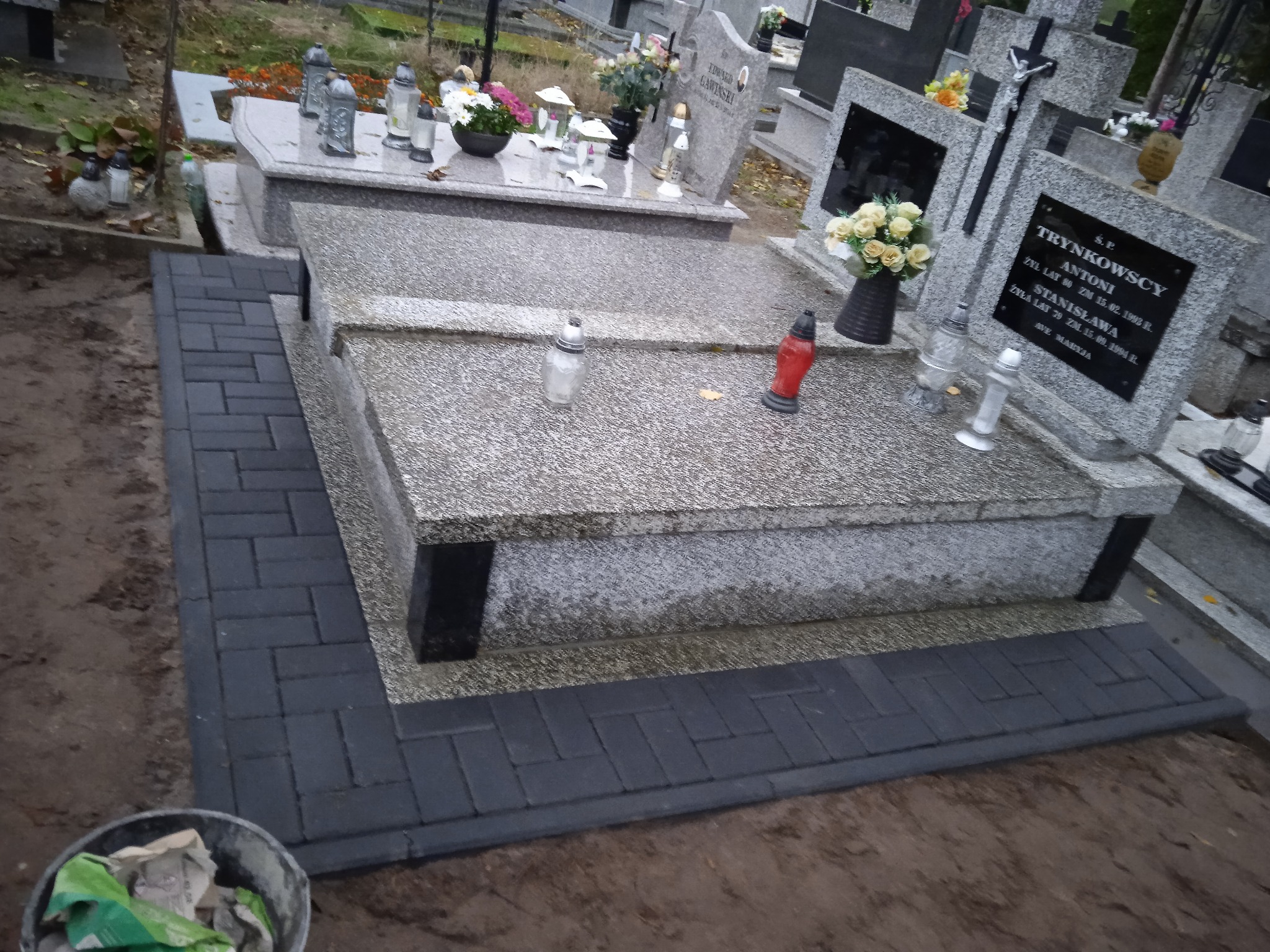 Ciemnoszara kostka brukowa ułożona wokół nagrobka z szarego kamienia na cmentarzu, widoczny fragment wiadra z materiałami budowlanymi.