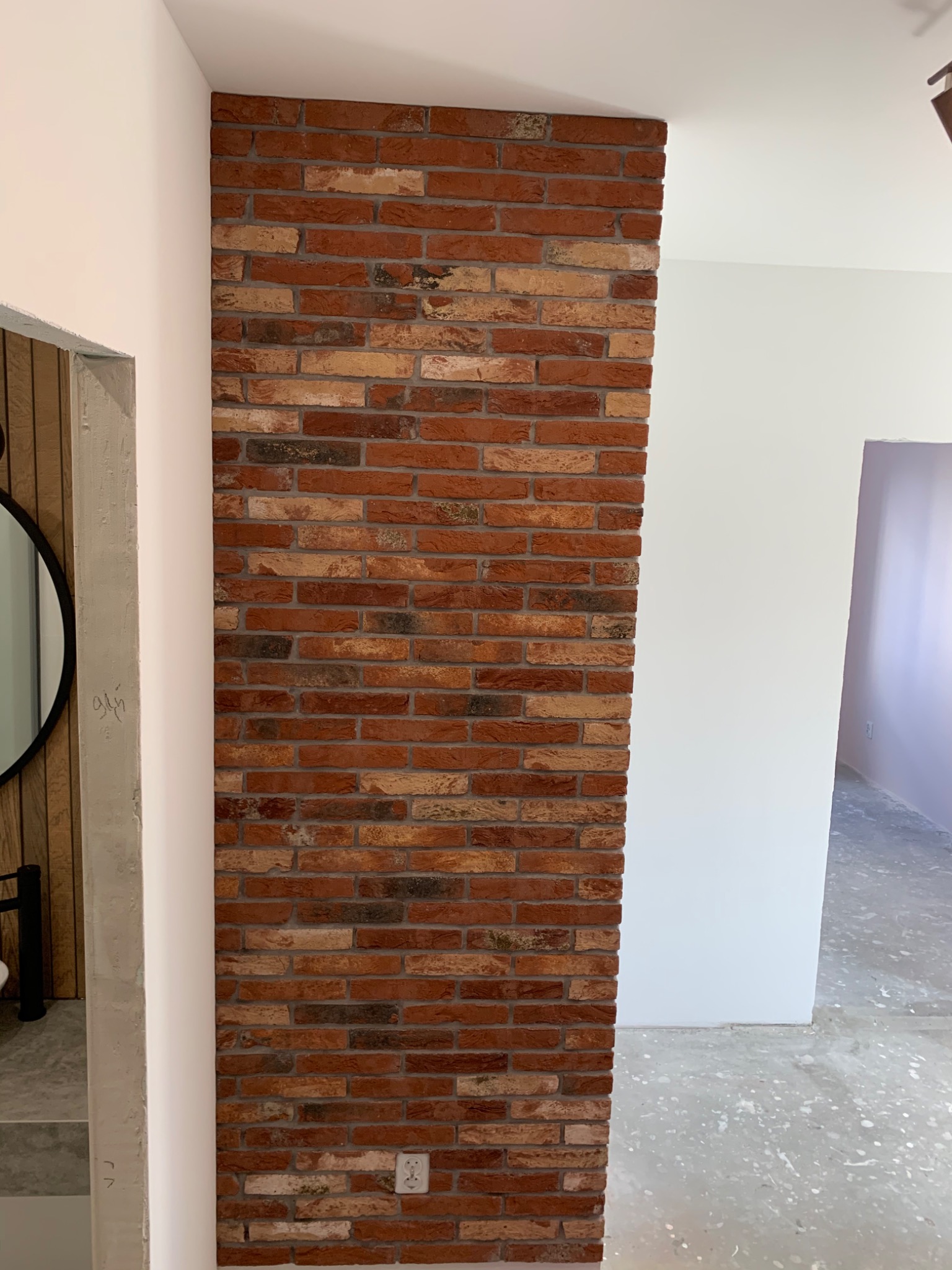 Fragment ściany wykończony czerwoną cegłą klinkierową, przylegający do białej ściany, z widocznym fragmentem ościeżnicy drzwiowej i częściowo widoczną podłogą w trakcie prac wykończeniowych.