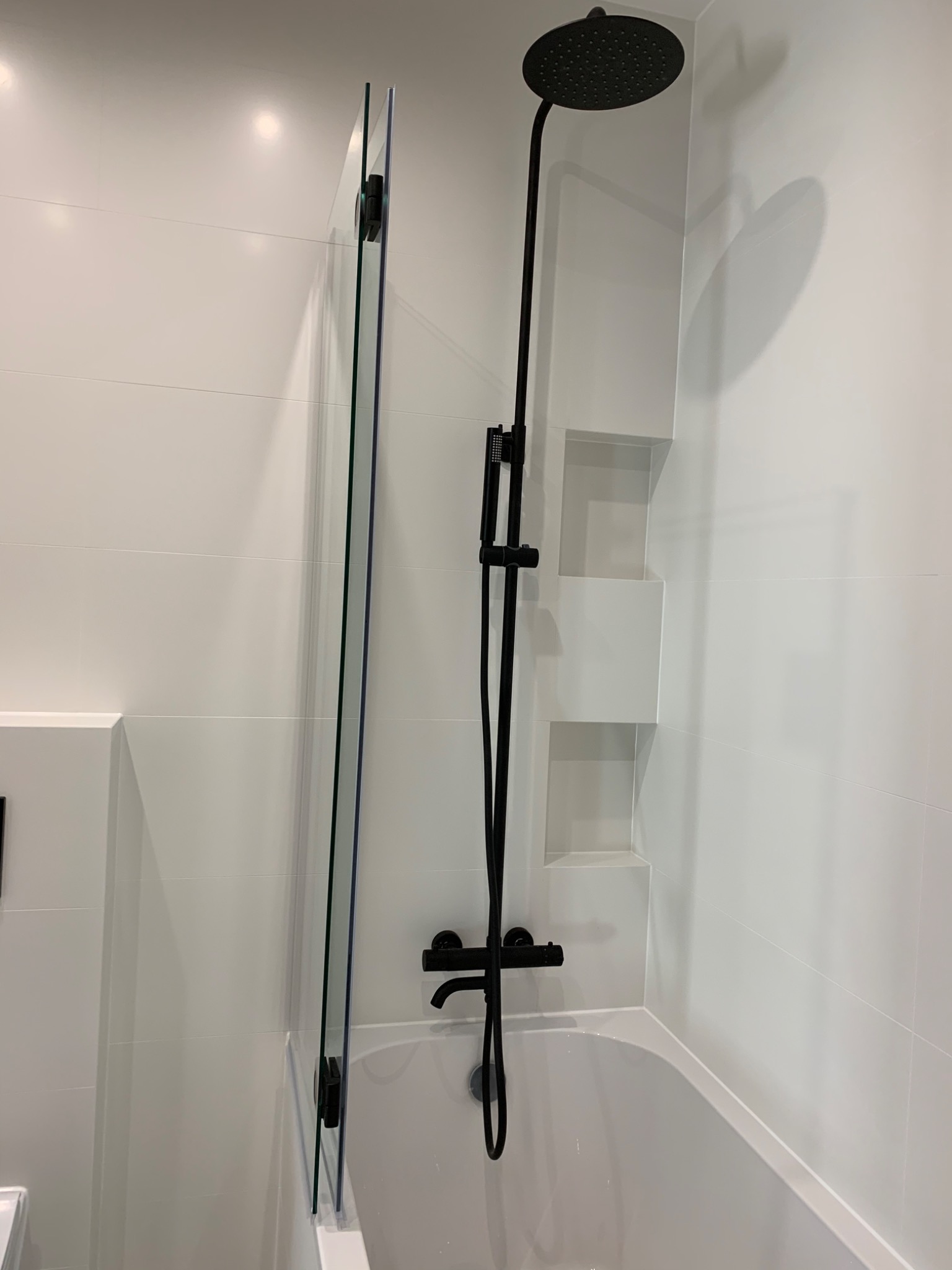 Nowoczesna łazienka z białą glazurą, czarną armaturą prysznicową i szklanym parawanem nawannowym.