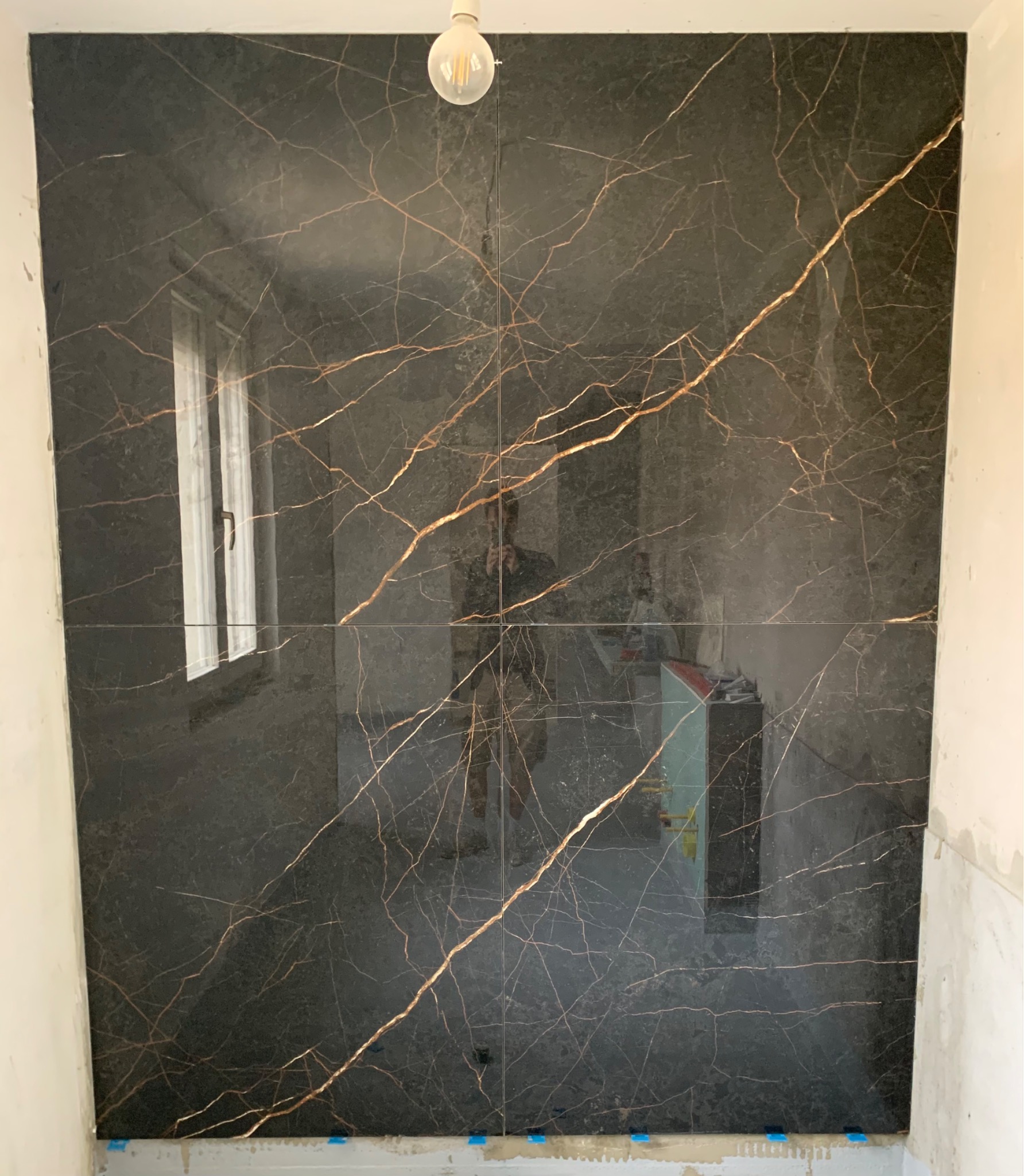 Ściana wyłożona czarnymi płytkami imitującymi marmur ze złotymi żyłami w łazience w Mikołowie, widoczne okno odbijające się w powierzchni płytek oraz robocza żarówka.
