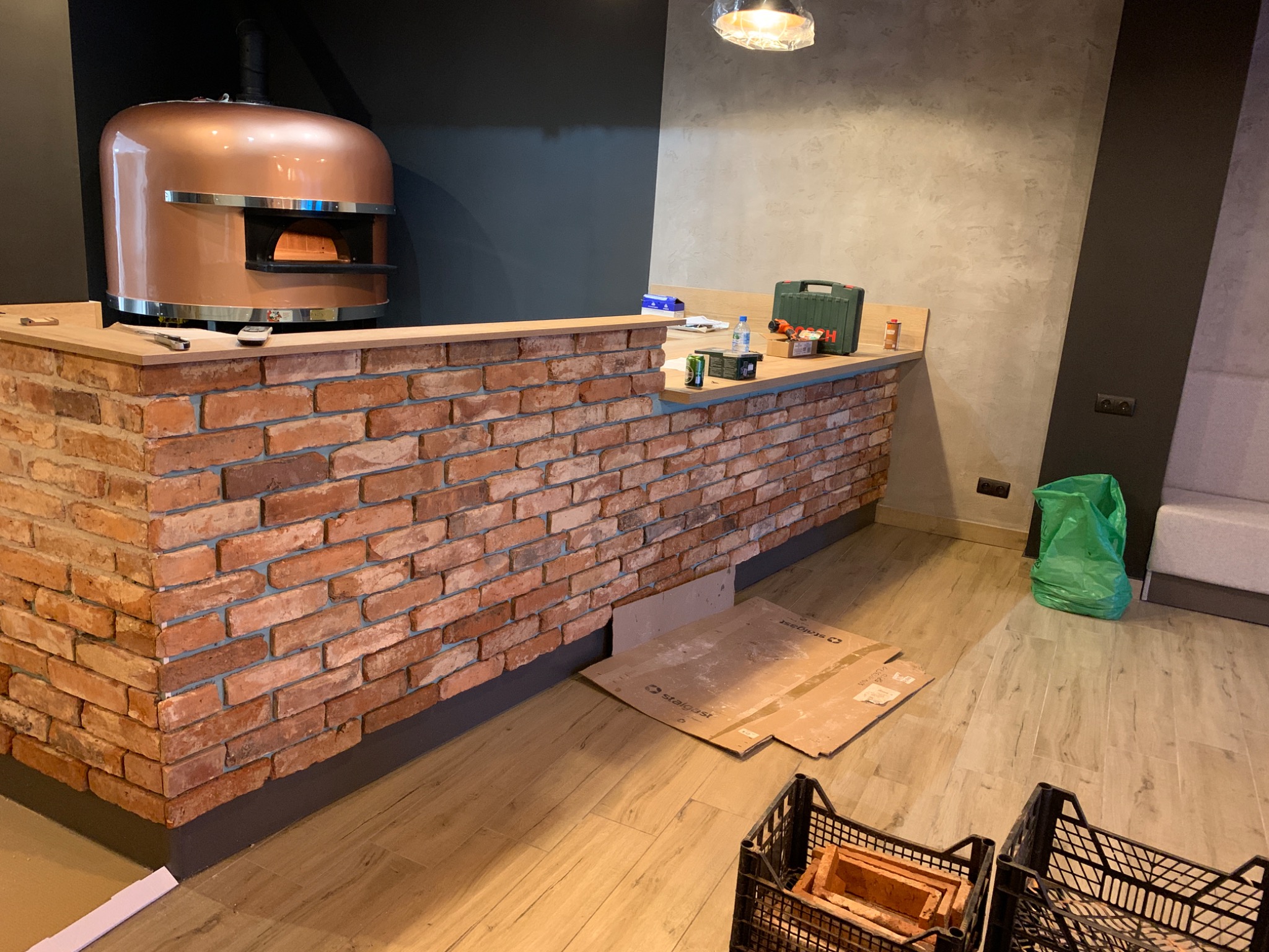 Wnętrze w trakcie wykańczania z ceglanym barem, na którym stoi miedziany piec do pizzy, widoczne narzędzia i materiały budowlane, podłoga wyłożona płytkami imitującymi drewno, częściowo...