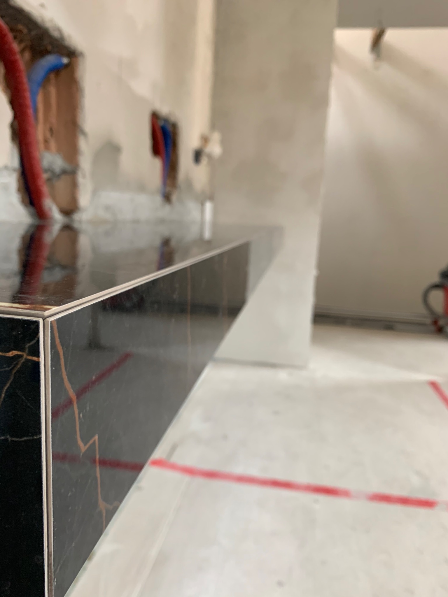 Czarny blat z żyłkowaniem w odcieniach złota, zamontowany w surowym wnętrzu łazienki, widoczne instalacje hydrauliczne i fragment odkurzacza przemysłowego w tle.