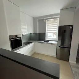 Nowoczesna, biała kuchnia z zabudową na wymiar, szary blat i metalowa lodówka, oświetlona naturalnym światłem z okna.