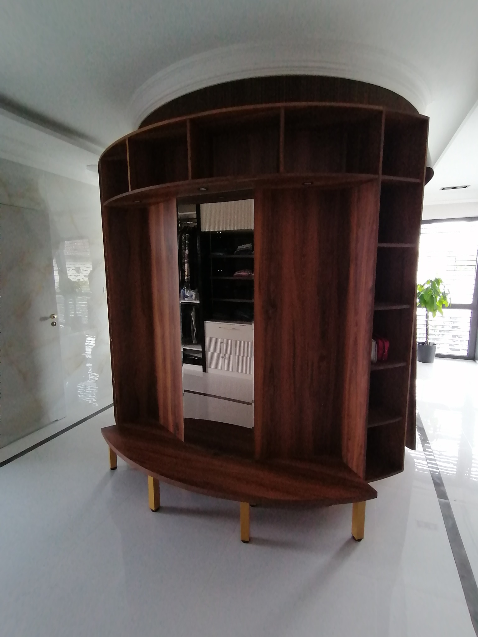 Zaokrąglona, wolnostojąca szafa z otwartymi półkami z ciemnego drewna na złotych nóżkach, w tle widoczna zawartość szafy wnękowej i roślina doniczkowa.