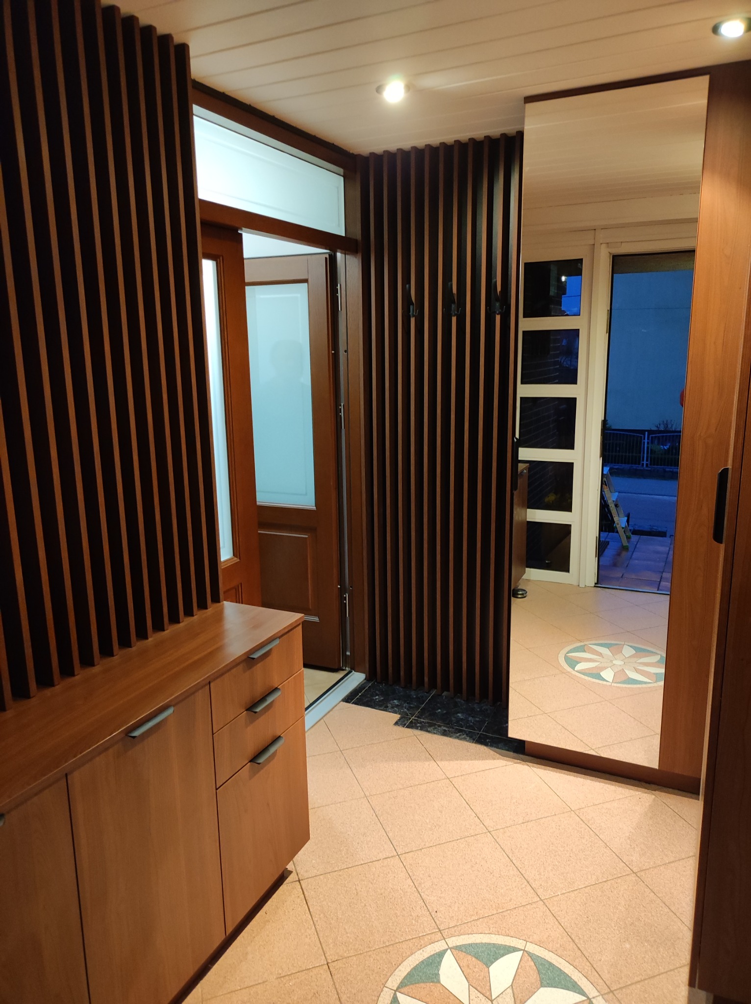 Wnętrze przedpokoju z drewnianą zabudową: szafka z szufladami, ażurowa ścianka z wieszakami i lustro w drewnianej ramie, widok na drzwi wejściowe z dekoracyjną szybą i wzorzystą posadzkę.