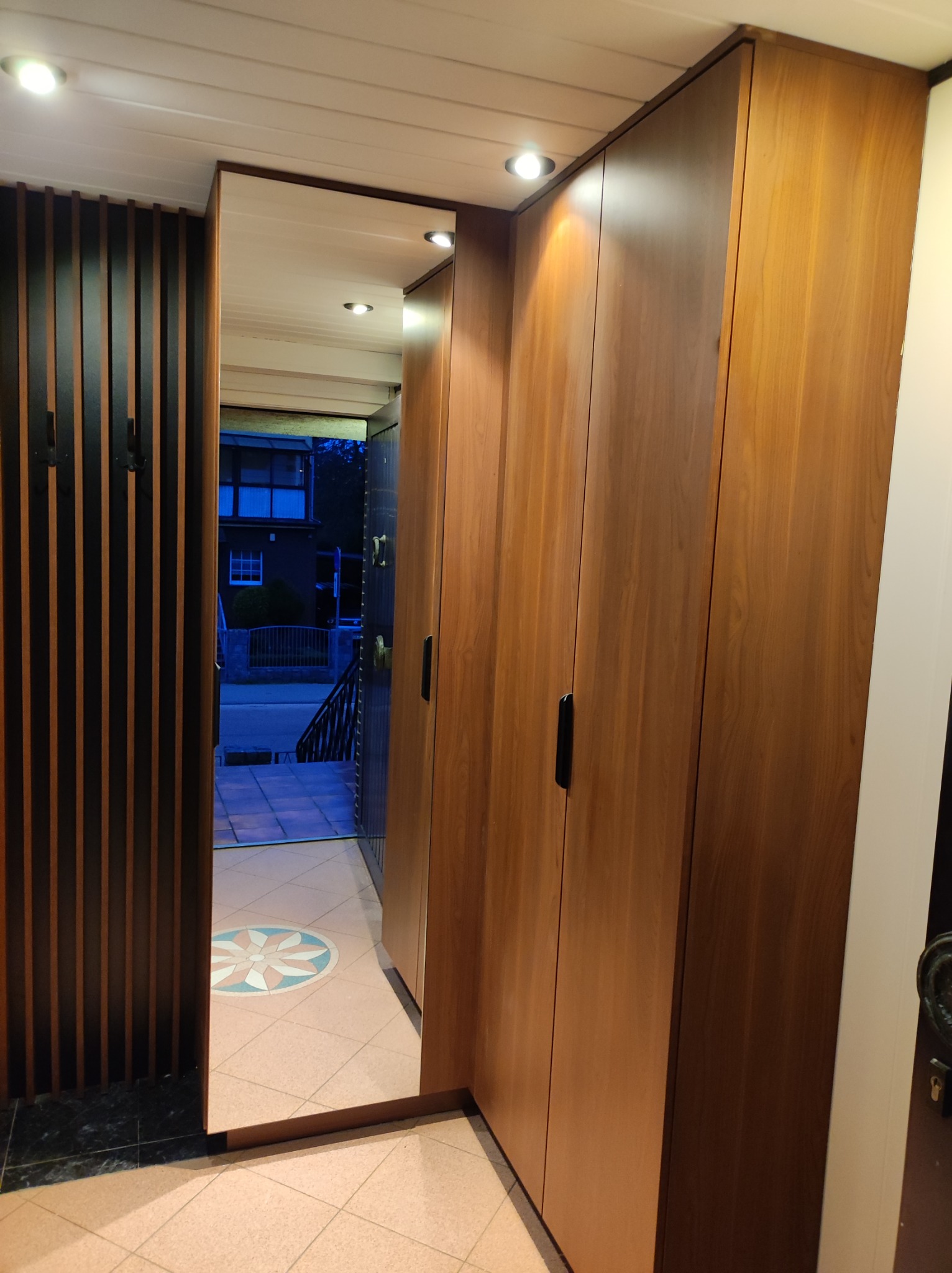 Wnętrze przedpokoju z zabudowaną szafą z drewnianymi frontami i dekoracyjną ścianką z pionowych listew, widok na zewnątrz przez otwarte drzwi.