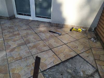 Demontaż brązowych płytek podłogowych na tarasie, widoczny fragment usuniętej płytki, narzędzia pracy: młotek i szlifierka kątowa.