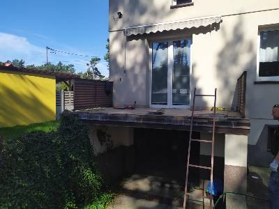 Zdjęcie przedstawia balkon w trakcie prac remontowych, widoczna drabina oparta o krawędź balkonu, częściowo usunięte płytki, zarośnięty dół balkonu, żółty budynek w tle.