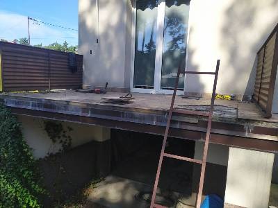 Demontaż starej podłogi na balkonie, widoczna drabina oparta o ścianę, narzędzia pracy i fragment elewacji budynku.