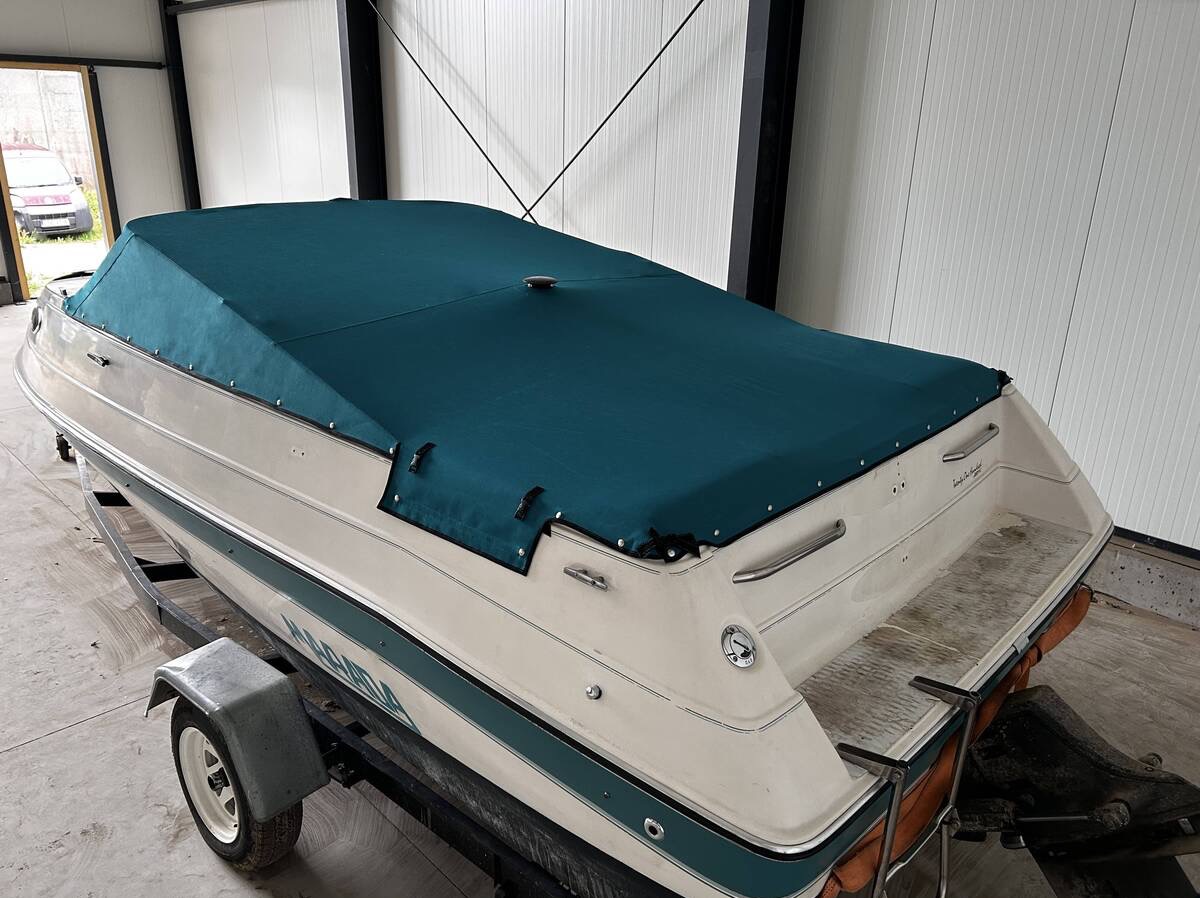 Łódź motorowa przykryta dopasowanym, ciemnozielonym pokrowcem z zatrzaskami, widoczna w jasnym pomieszczeniu z blachy falistej.