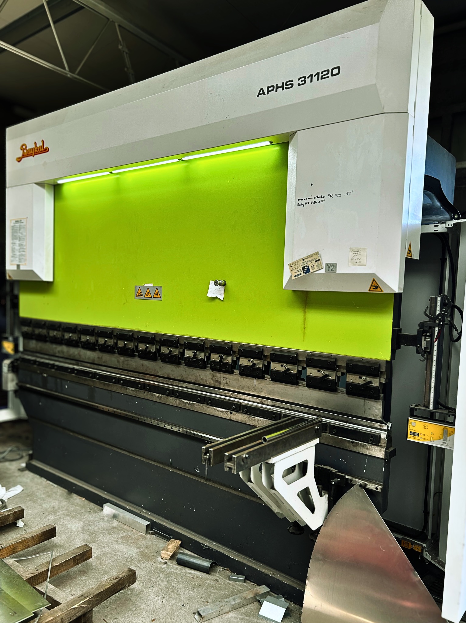 Duża, przemysłowa prasa krawędziowa marki Baykal, model APHS 31120, z zielonym panelem i widocznymi elementami sterującymi. Wokół porozrzucane metalowe elementy.