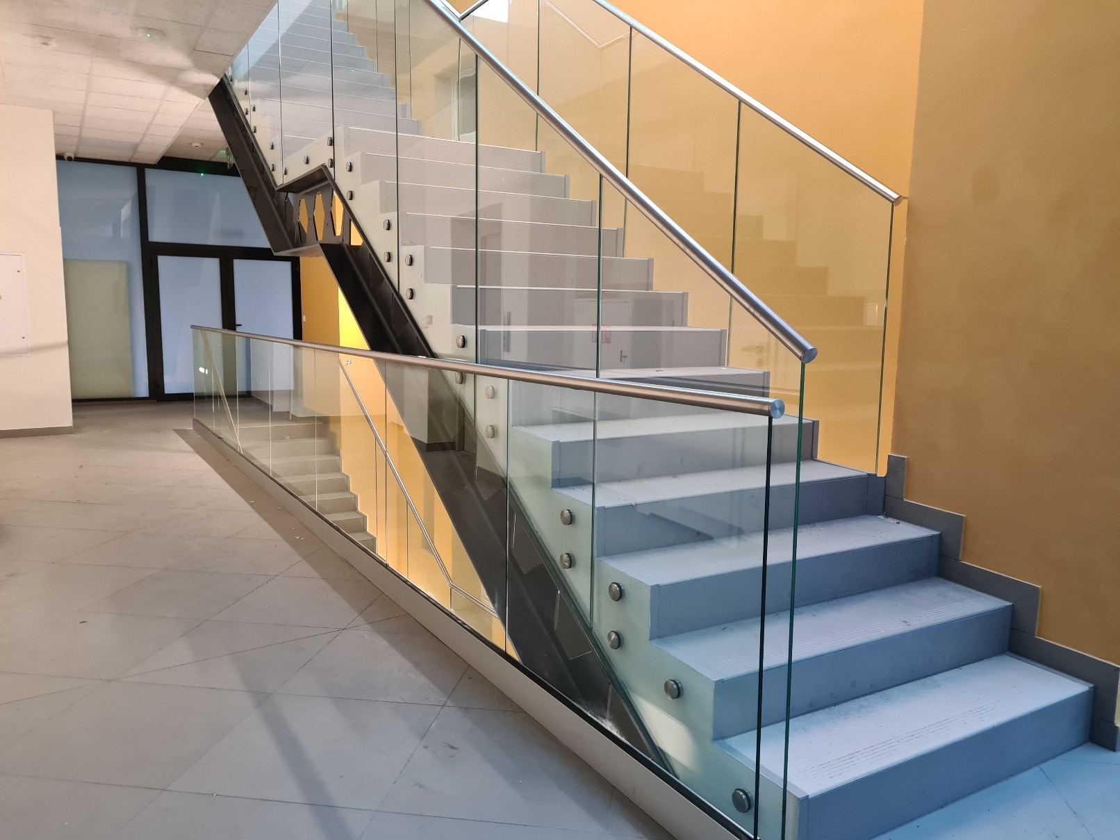 Nowoczesne schody wewnętrzne z szarymi stopniami, balustradą ze stali nierdzewnej i szkła, widoczne mocowania punktowe szkła, w tle pomarańczowa ściana.