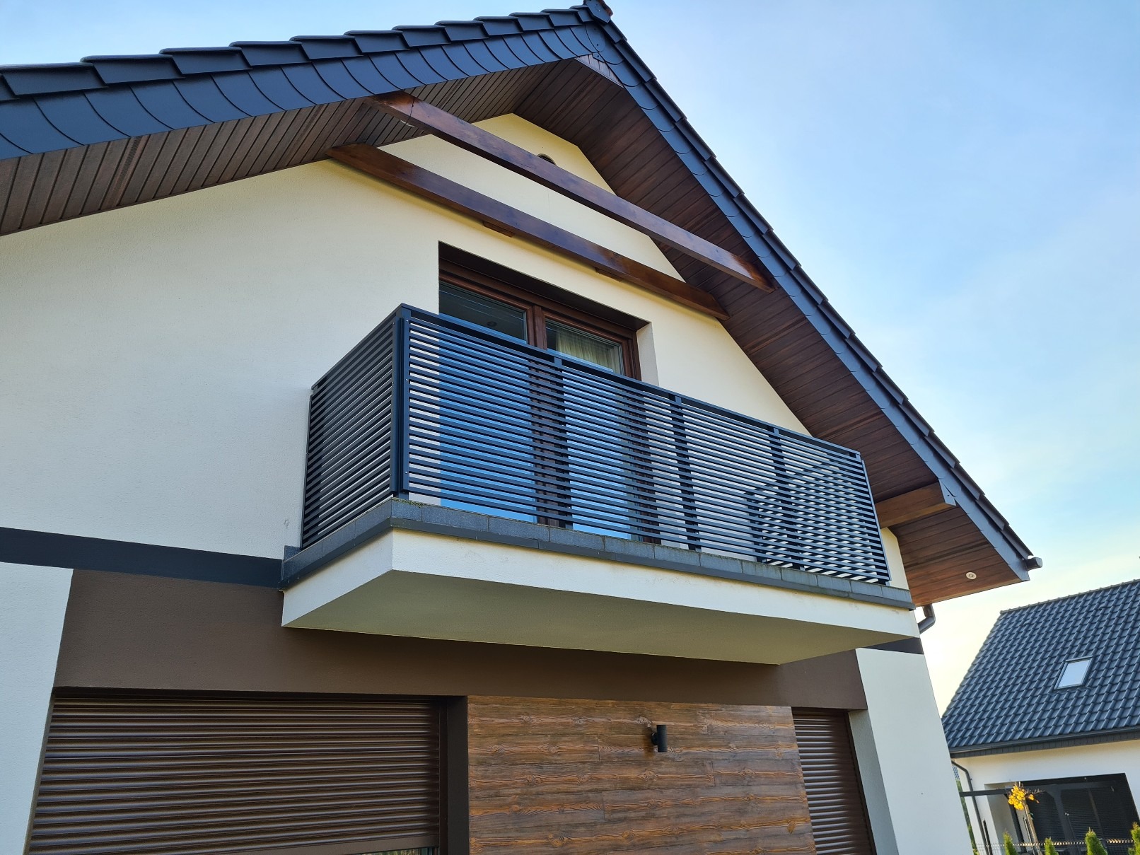Nowoczesna balustrada balkonowa z poziomych profili, zamontowana w domu jednorodzinnym z ciemną dachówką i drewnianymi elementami wykończeniowymi.