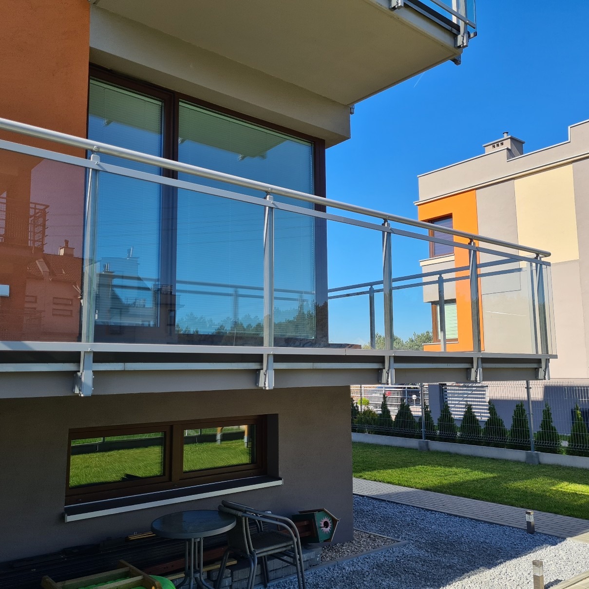 Nowoczesna balustrada szklana z elementami metalowymi na balkonie w Bydgoszczy, widok z dołu, z odbiciem trawnika w oknach piwnicznych.