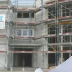 Inżynier w kasku z logo na tle budynku w trakcie budowy, trzymający plany.