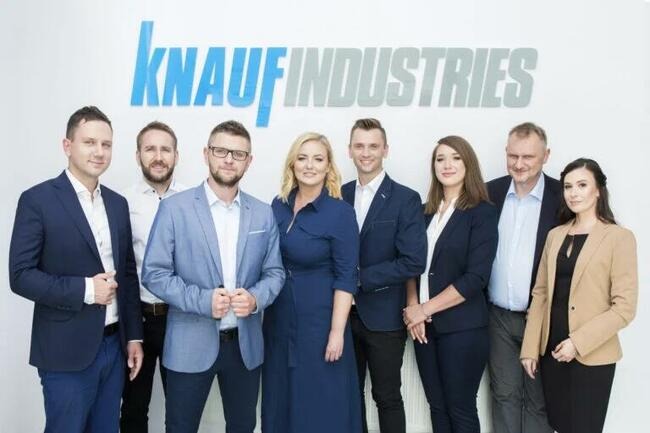 Grupa ośmiu osób w garniturach i strojach biznesowych, stojąca przed białym tłem z logo Knauf Industries w kolorze niebieskim i szarym.