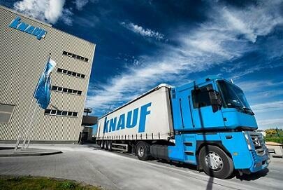 Ciężarówka z naczepą z logo Knauf na tle budynku fabryki Knauf z powiewającą flagą, błękitne niebo z chmurami