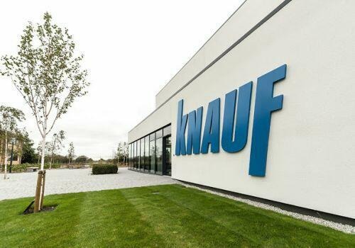 Budynek z dużym, niebieskim logo Knauf na białej fasadzie, otoczony trawnikiem i drzewami.