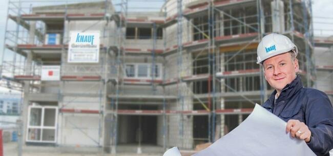 Inżynier w kasku z logo na tle budynku w trakcie budowy, trzymający plany.