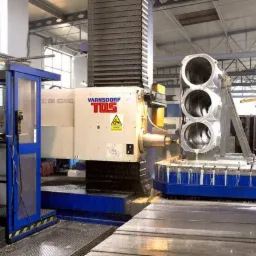 Operator w niebieskim kombinezonie obsługuje frezarkę CNC VARNSDORF TOS podczas obróbki metalowego elementu z trzema otworami, chłodziwo spływa po obrabianym przedmiocie, w tle widoczna hala...