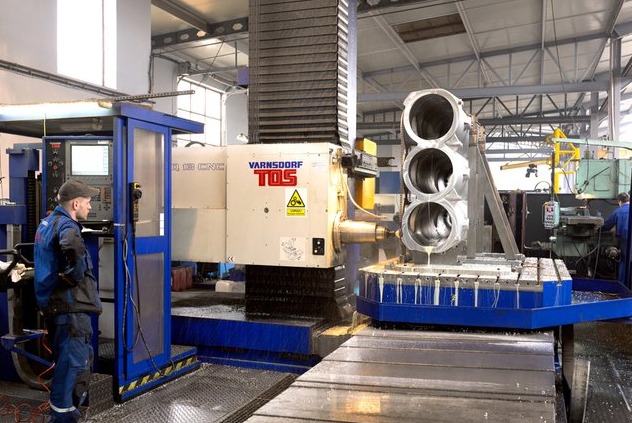 Operator w niebieskim kombinezonie obsługuje frezarkę CNC VARNSDORF TOS podczas obróbki metalowego elementu z trzema otworami, chłodziwo spływa po obrabianym przedmiocie, w tle widoczna hala...