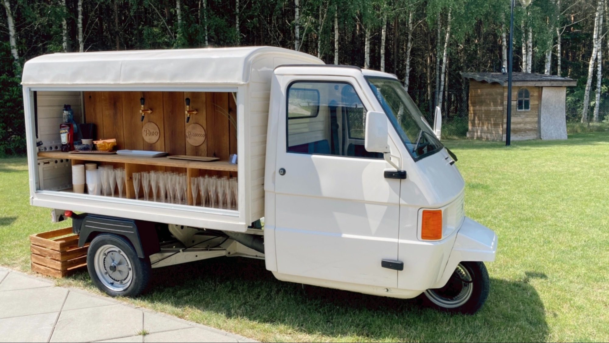 Mobilny bar w białym samochodzie trójkołowym z drewnianym blatem, kranami do piwa i prosecco, szklankami, butelką Aperolu i przekąskami, ustawiony na trawie przed lasem.