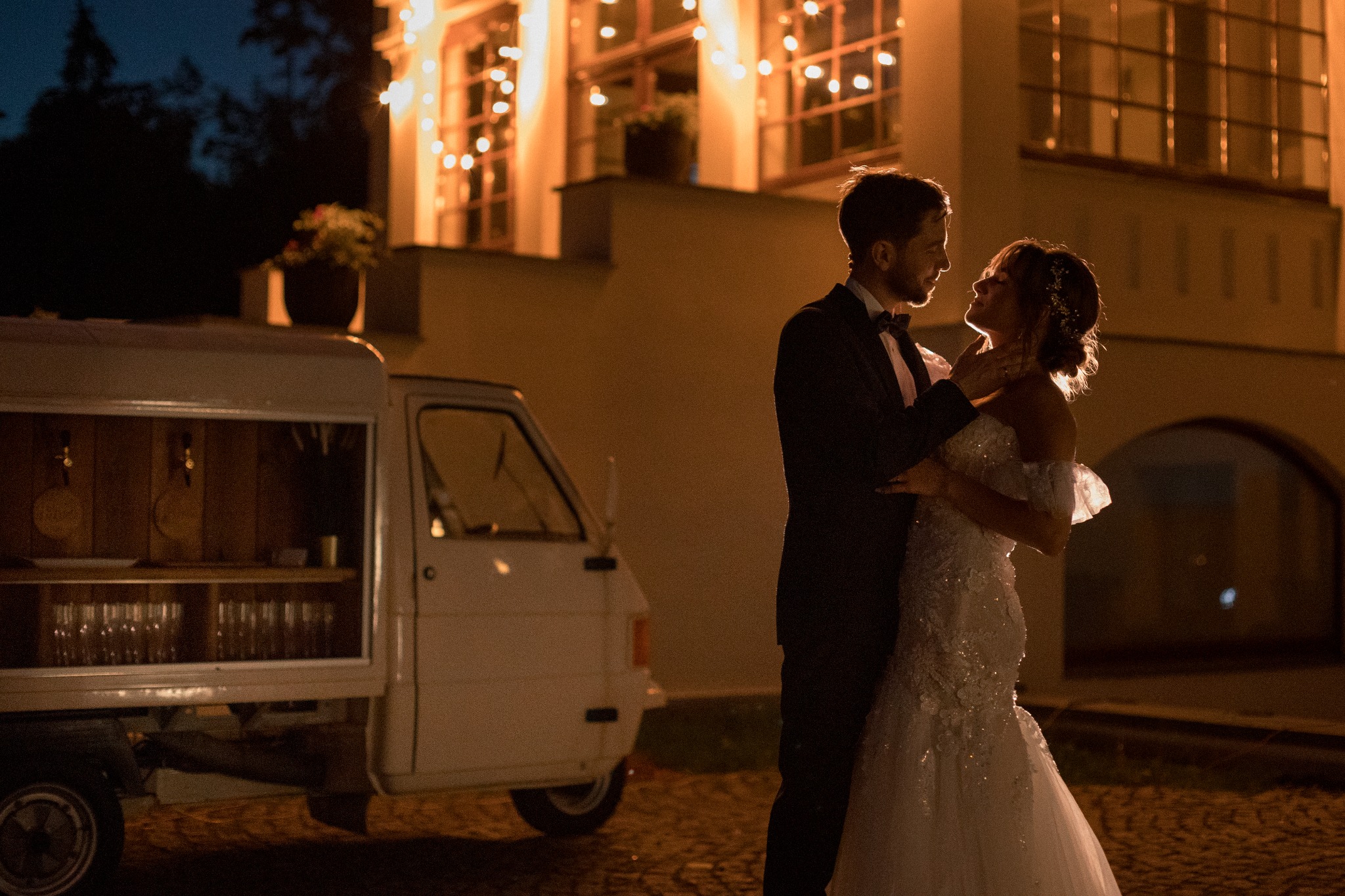 Para młoda w objęciach na tle budynku z oknami oświetlonymi girlandami i mobilnego baru z zapasem szkła, wieczorna sceneria.
