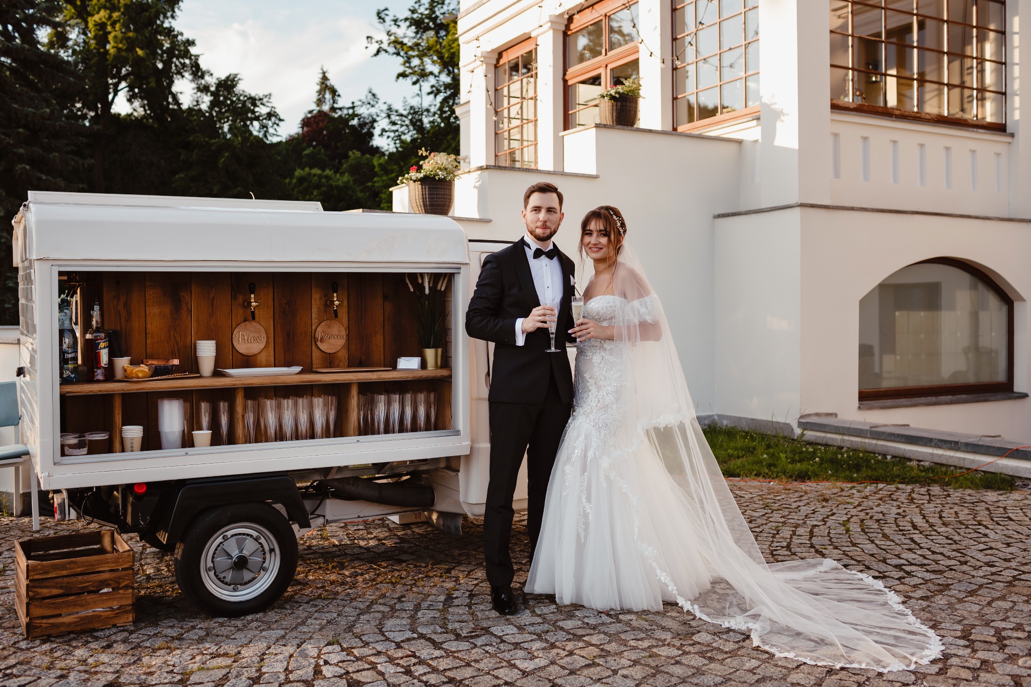 Para młoda w strojach ślubnych pozuje z kieliszkami szampana obok mobilnego baru z drewnianym wykończeniem, z kranami do piwa i prosecco, na tle białego budynku z dużymi oknami.