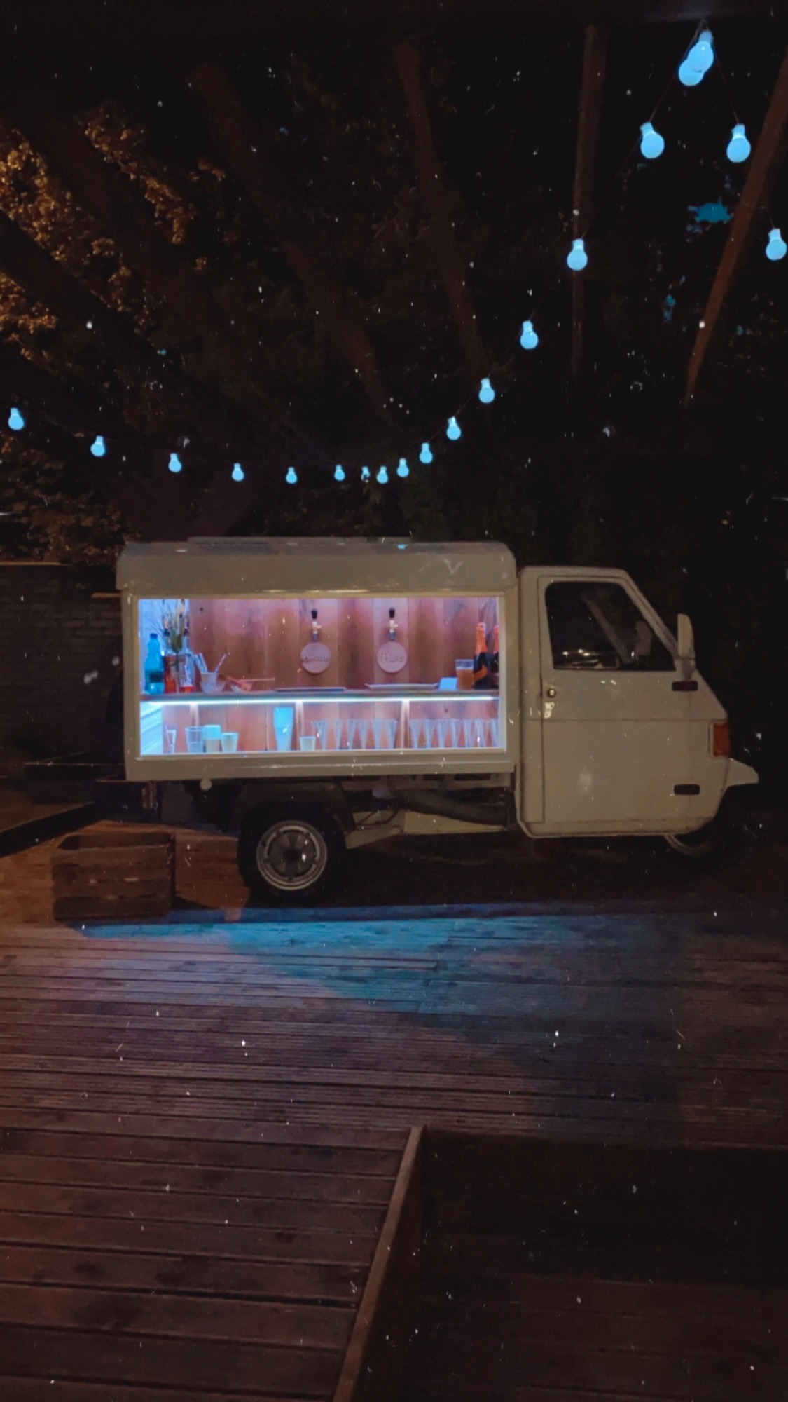 Biały mobilny bar w samochodzie Piaggio Ape na drewnianym podeście, oświetlony girlandą żarówek, przygotowany do serwowania napojów.