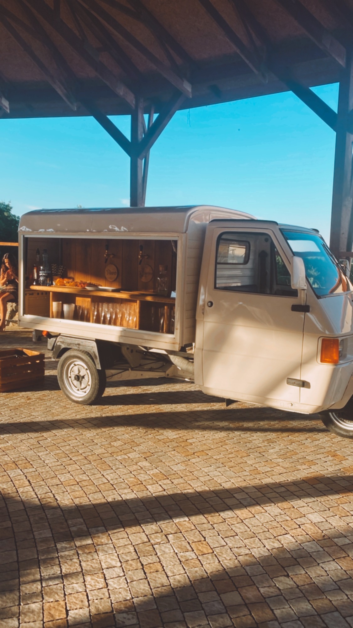 Mobilny bar w vanie, wyposażony w szklanki, napoje i przekąski, ustawiony na brukowanym placu pod zadaszeniem, z osobami w tle.
