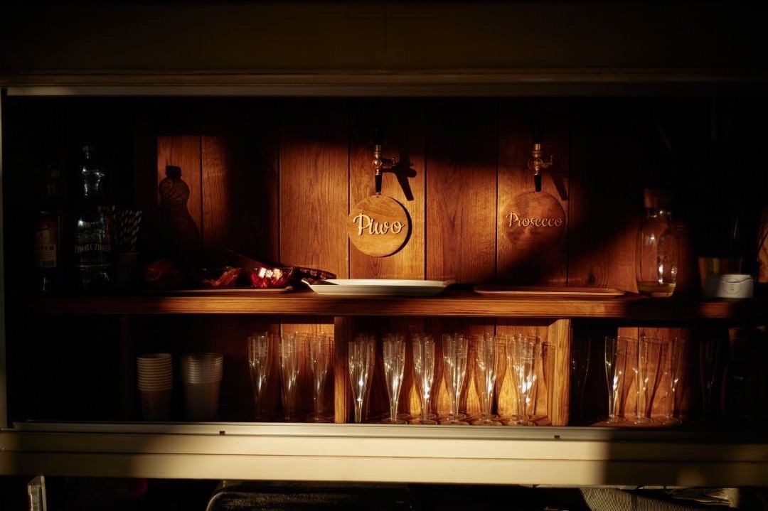 Drewniany bar z kranami do piwa i prosecco, szklanki do szampana ustawione w rzędzie, oświetlenie punktowe podkreśla napisy na kranach.
