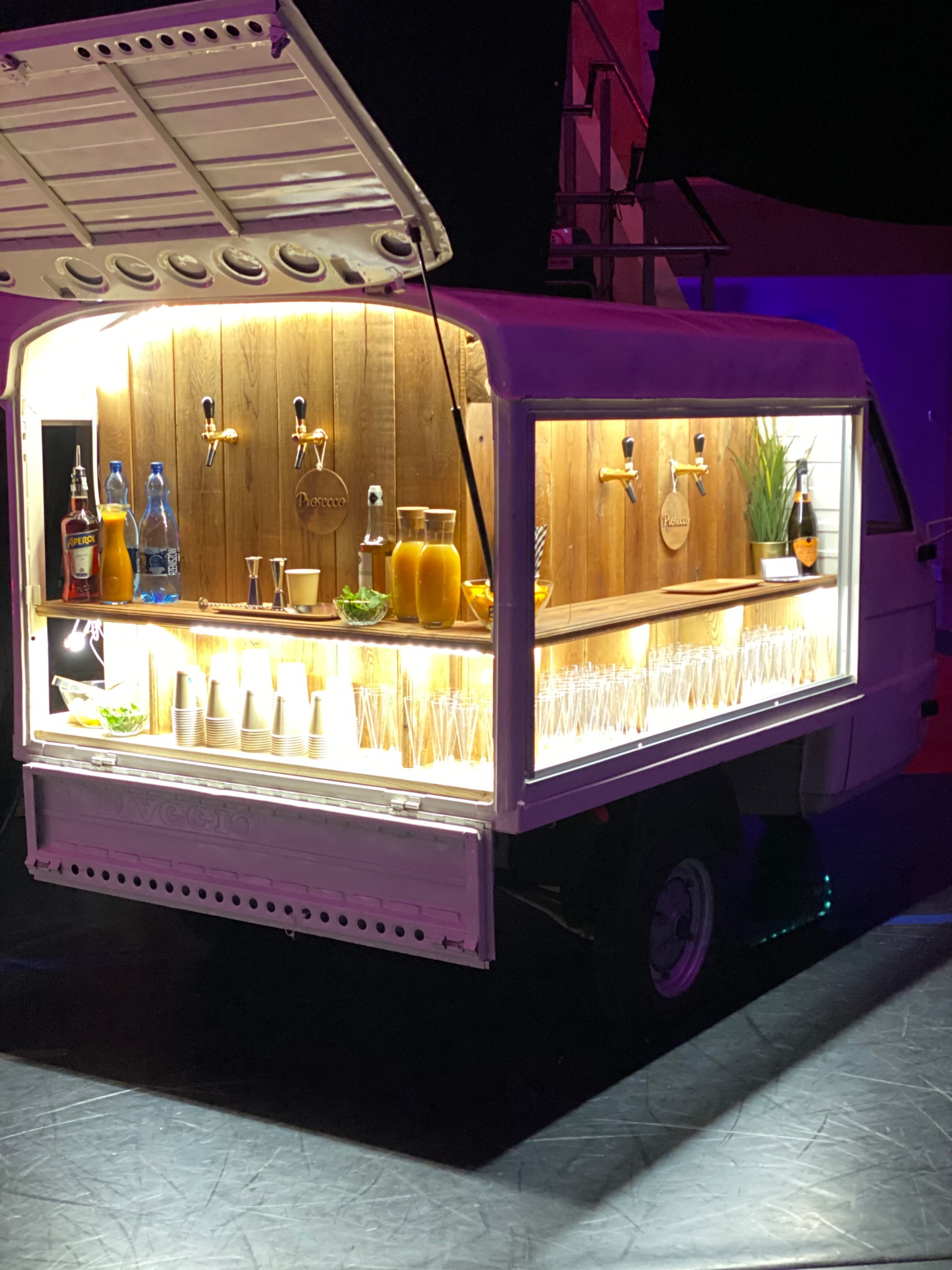 Mobilny bar w przyczepie Ape Piaggio, oświetlony od wewnątrz, z drewnianym blatem, kranami do napojów, butelkami, szklankami i dekoracjami roślinnymi.