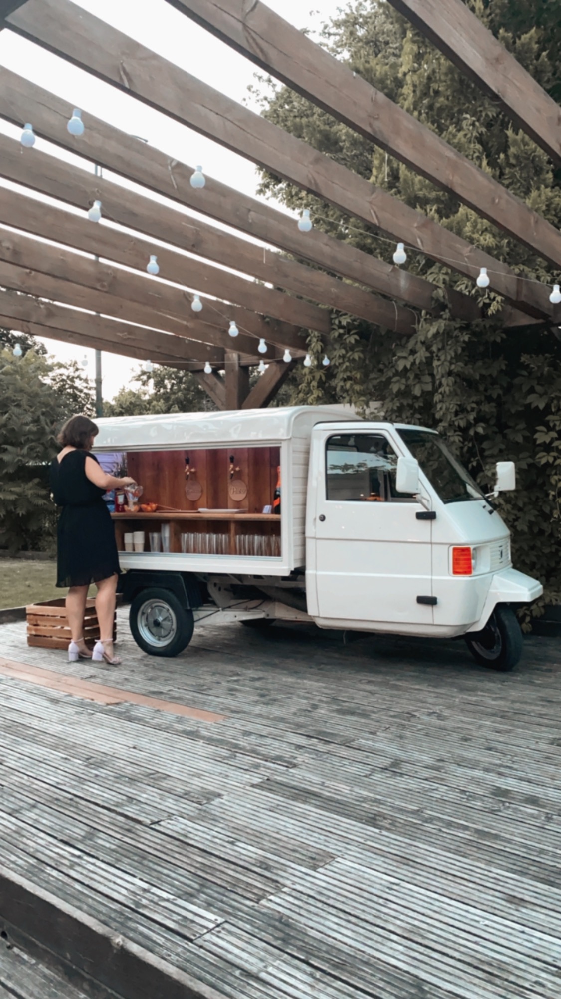 Mobilny bar na bazie białego trójkołowca Ape, obsługiwany przez kobietę w czarnej sukience, na drewnianym podeście, oświetlony girlandami żarówek.