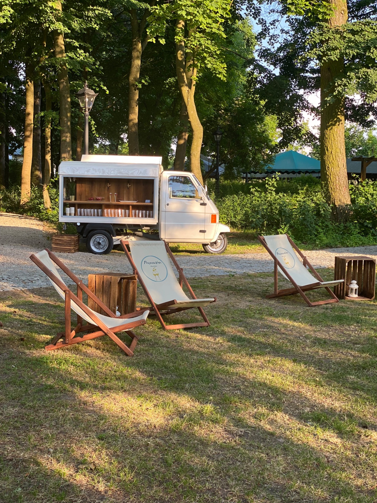 Biały food truck z otwartą przestrzenią barową, ustawiony na trawie w otoczeniu drzew, z trzema leżakami z logo i drewnianymi skrzynkami, tworząc strefę relaksu na imprezie plenerowej.