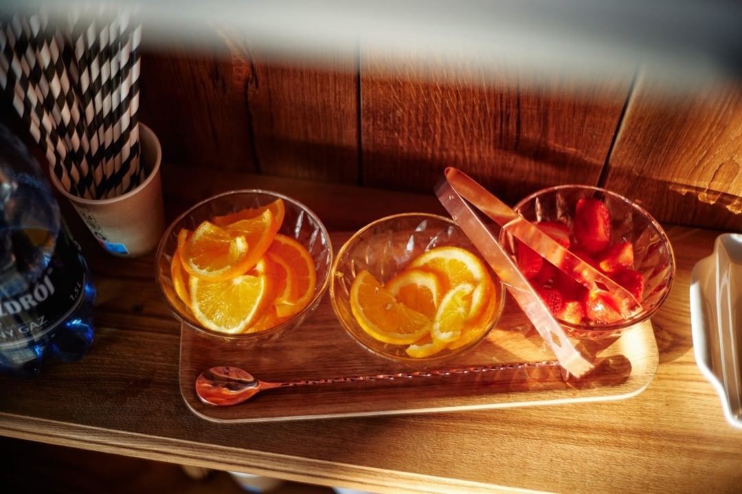 Przygotowane na drewnianej tacy szklane miseczki z pokrojonymi pomarańczami i truskawkami, obok szczypce, długa łyżka barmańska, butelka wody i kubek ze słomkami, oświetlone ciepłym światłem.