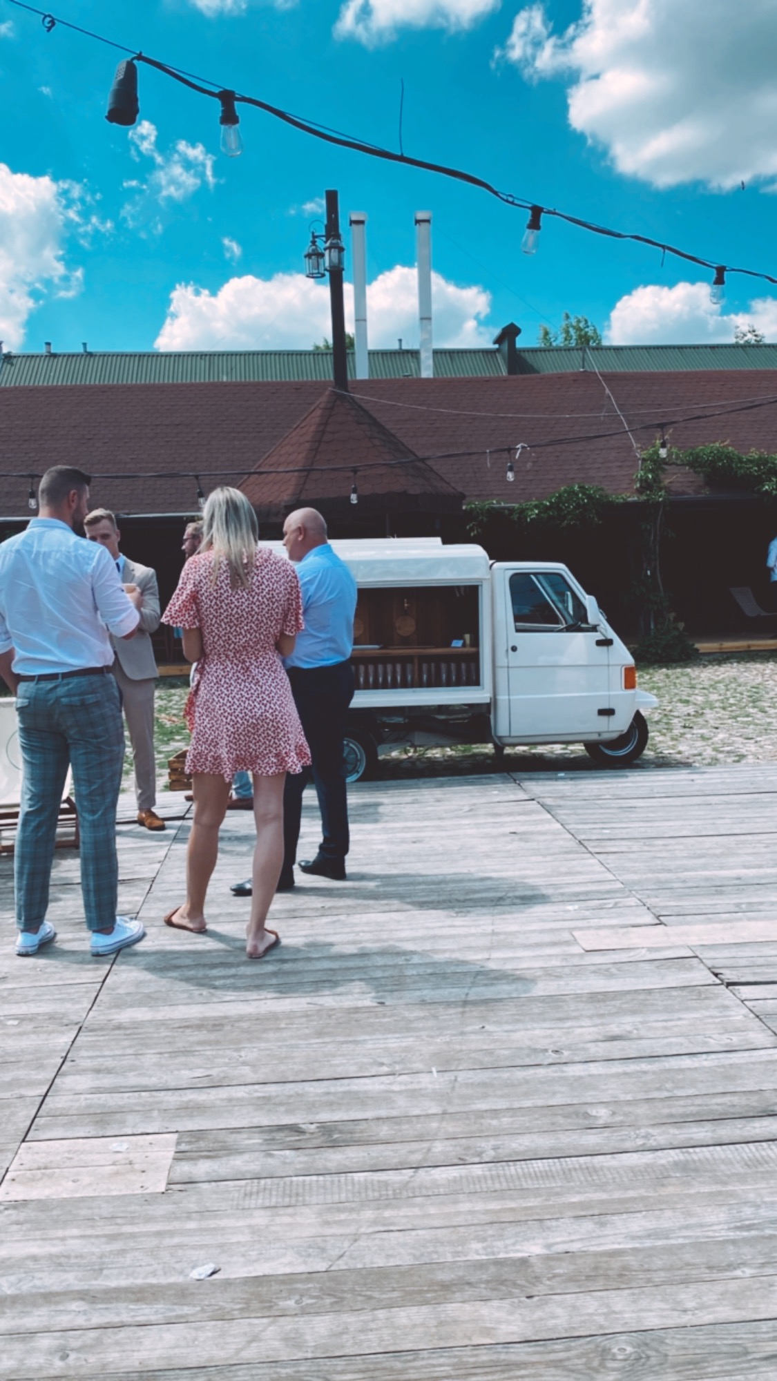 Mobilny bar na bazie trójkołowego pojazdu, obsługujący grupę osób podczas imprezy plenerowej na drewnianym podeście, z dekoracyjnym oświetleniem i budynkiem w tle.