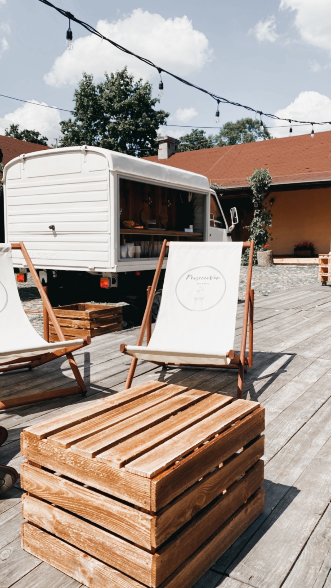 Mobilny bar ProseccoVan z dwoma leżakami z logo i stolikiem z drewnianych skrzynek na drewnianym tarasie, girlanda żarówek w tle.