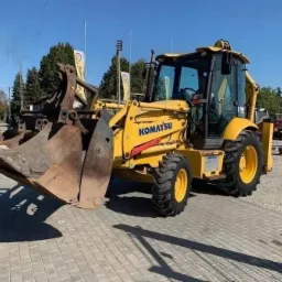 Żółta koparko-ładowarka Komatsu z łyżką i chwytakiem na placu w Górze Kalwarii, widoczna na tle błękitnego nieba i drzew.