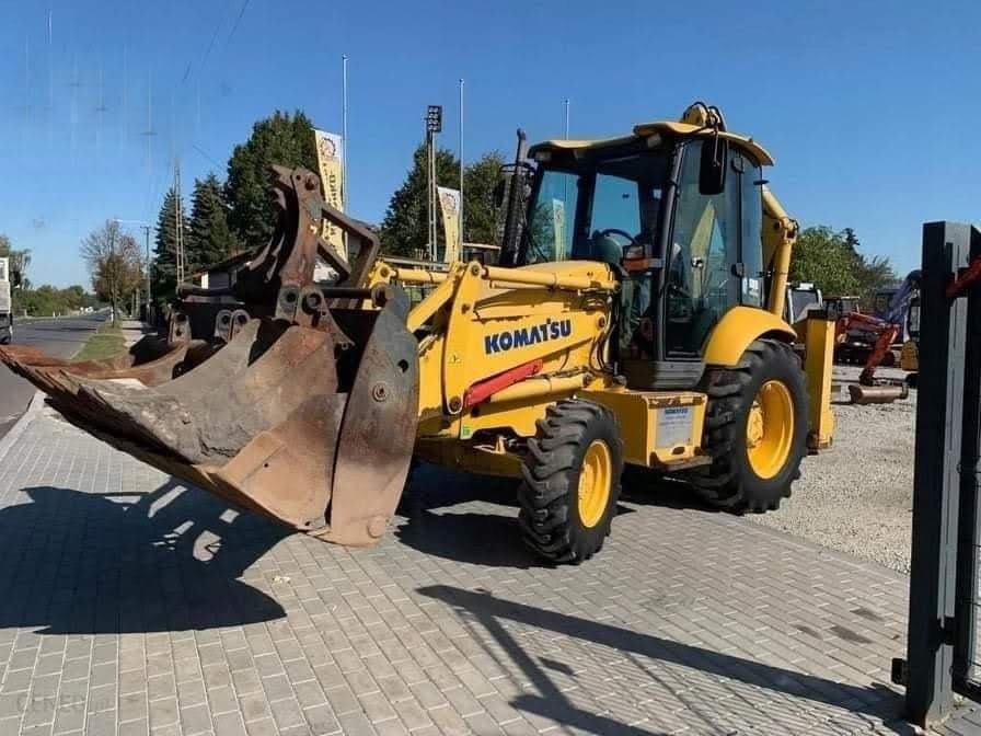 Żółta koparko-ładowarka Komatsu z łyżką i chwytakiem na placu w Górze Kalwarii, widoczna na tle błękitnego nieba i drzew.